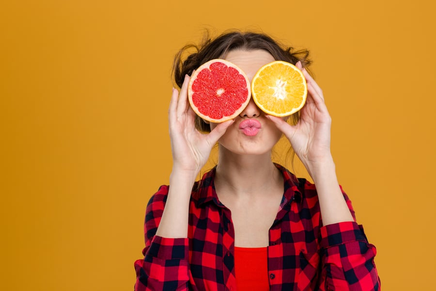 Funny playful young woman in checkered shirt holding halves of citrus fruits against her eyes and making duck face over yellow background