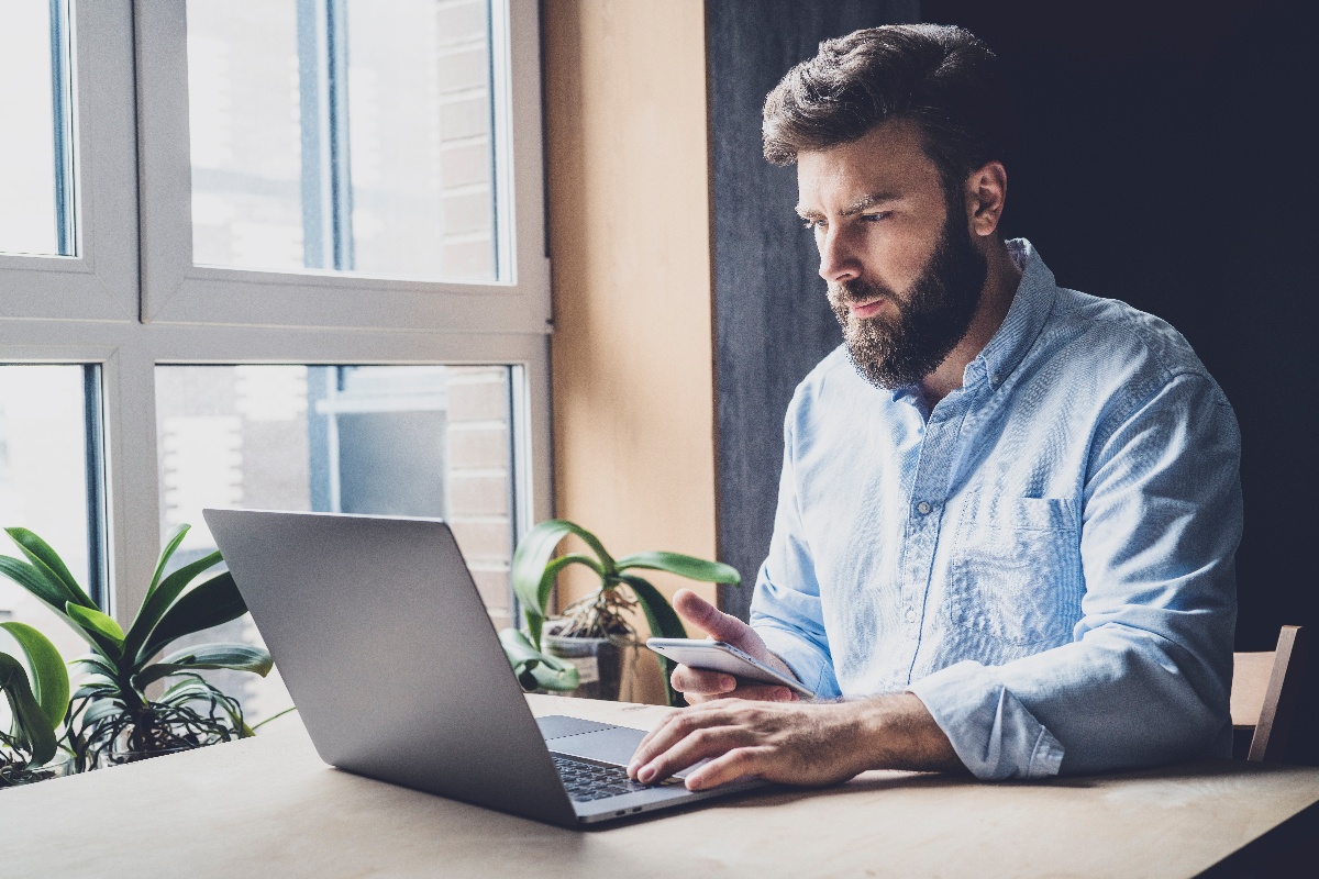 Man working by window in office using wireless digital gadgets for projects. Young professional checking email and sending corporate letters. Office manager typing on laptop and communicating on phone-1